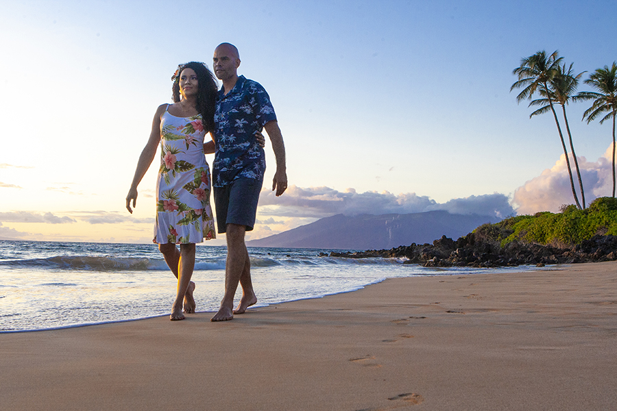 A couple walks barefoot along a tropical beach at sunset, with palm trees and distant mountains in the background.