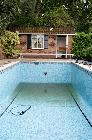 Empty outdoor swimming pool with blue mosaic tiles, adjacent brick pool house, and garden surroundings.