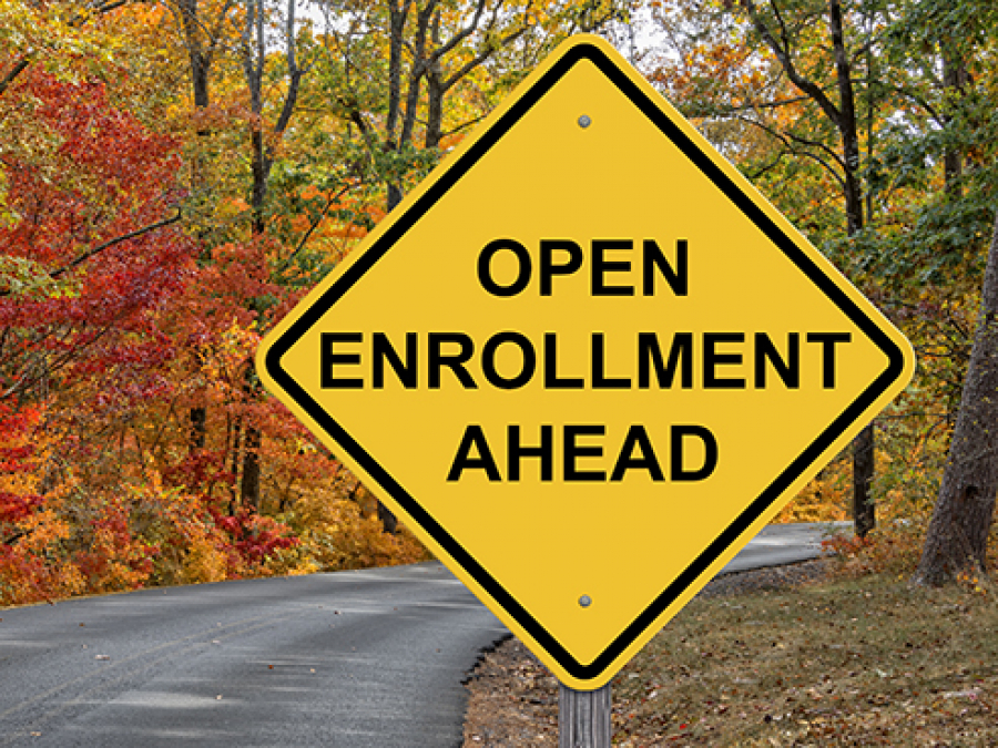 Yellow diamond-shaped road sign reading "OPEN ENROLLMENT AHEAD" against autumn forest backdrop.