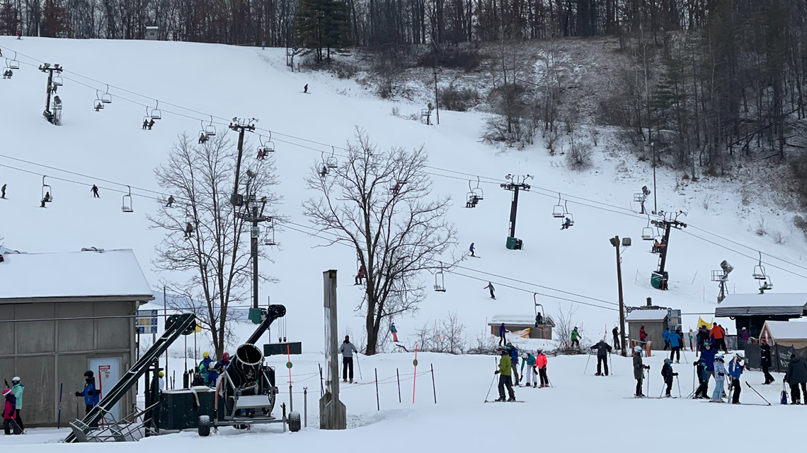 Skiers and snowboarders on a snowy slope with chairlifts, snow cannon, and bare trees in winter at a ski resort.