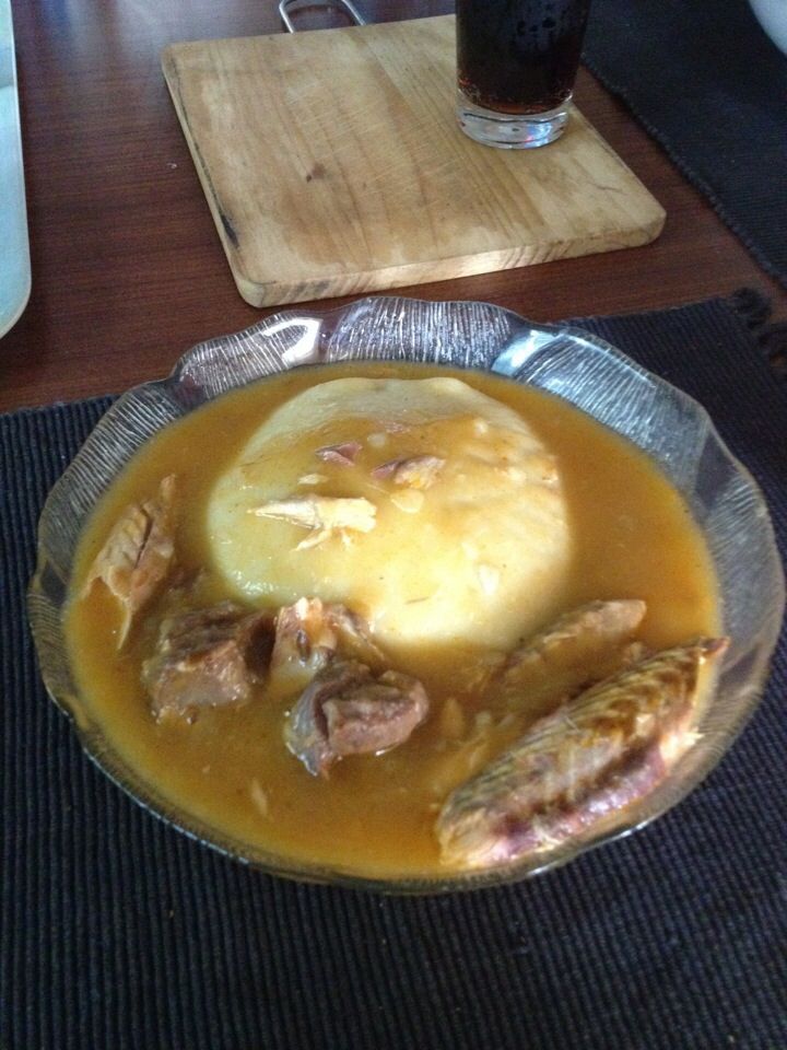 A bowl of traditional stew with fish, meat, and a mound of fufu or pounded yam in rich broth, served on a dark placemat.