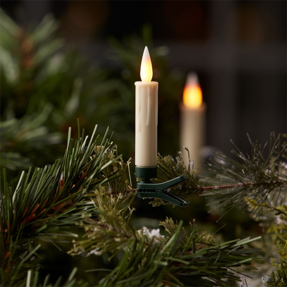 LED flameless candle clipped to pine branch, warm glow among green needles and snowy accents.