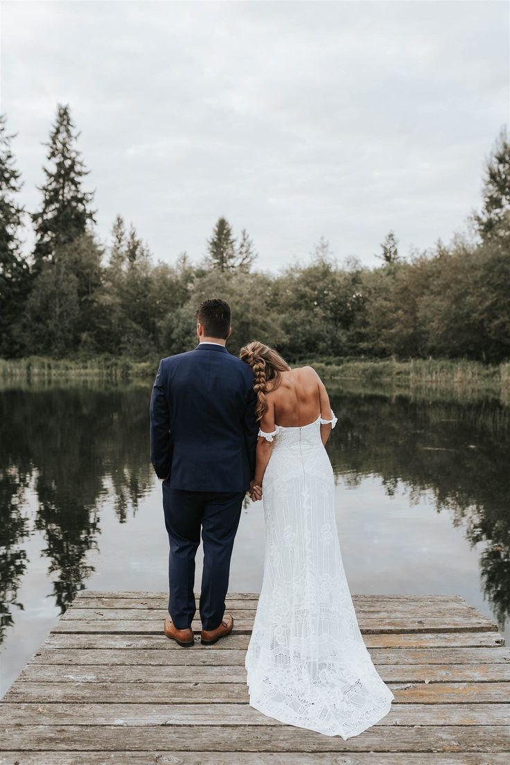 A bride and groom stand hand-in-hand on a wooden dock, gazing at a serene forest-lined lake under an overcast sky.