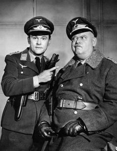 Two actors in WWII-era German military uniforms, one pointing a pistol at the other in a comedic pose.