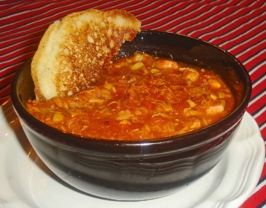 Hearty tomato-based stew with shredded chicken and corn, served in a dark ceramic bowl with toasted bread on a red-striped tablecloth.