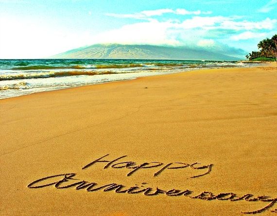 "Happy Anniversary" written in sand on a tropical beach with ocean and mountain backdrop.