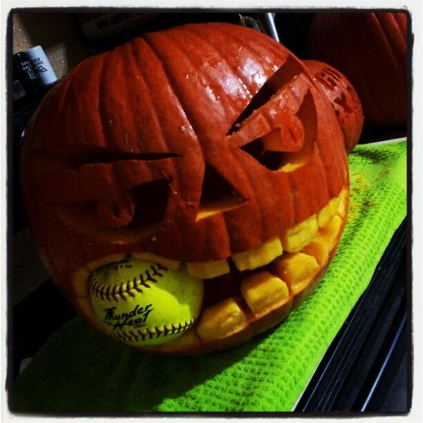 Carved pumpkin with angry face holding a yellow "Thunder Heat" softball in its mouth, resting on a green towel.
