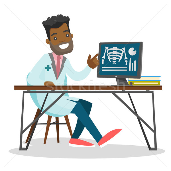 Smiling Black male doctor in lab coat pointing at X-ray of ribcage on computer monitor while seated at desk.