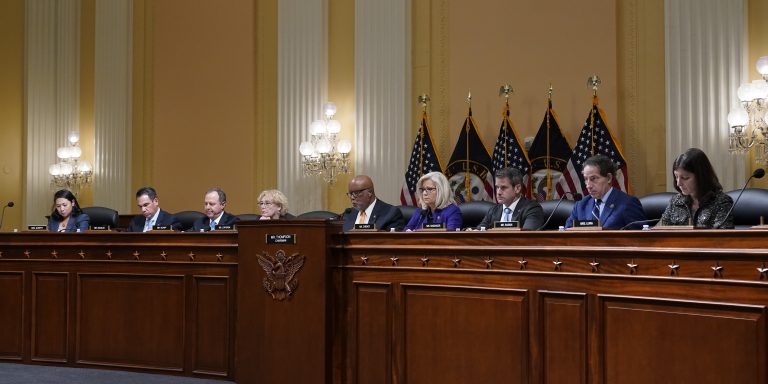 U.S. congressional committee members seated at a formal hearing table with flags and ornate chamber background.