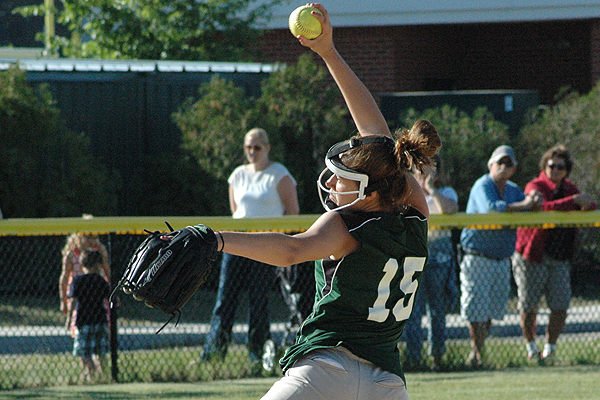 Female softball pitcher in green jersey #15 mid-throw, wearing helmet and glove, with spectators behind fence.
