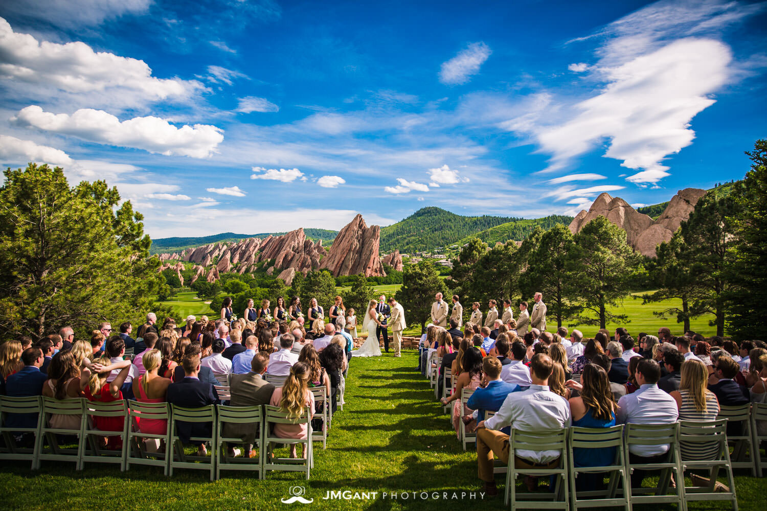 Outdoor wedding ceremony at Garden of the Gods with red rock formations and guests seated on lawn under blue sky.