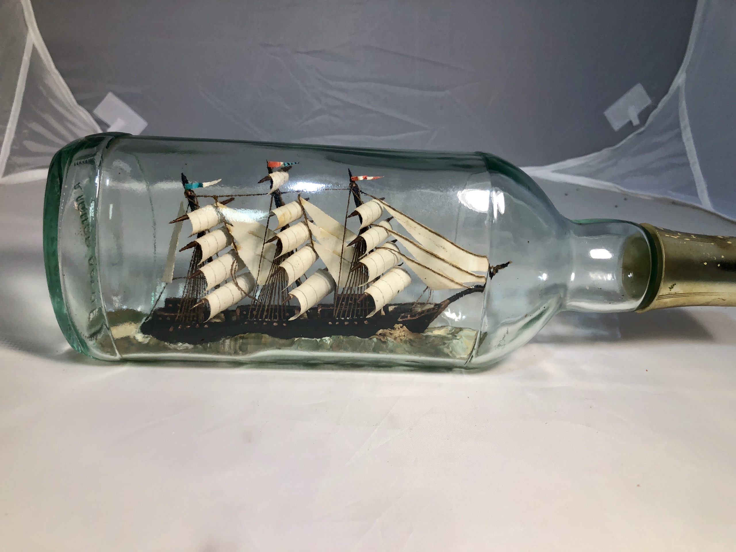 Vintage ship-in-a-bottle model with detailed sails and rigging inside a green-tinted glass bottle, lying horizontally on white fabric.