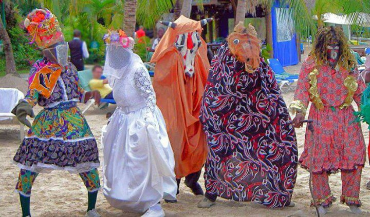 Colorful traditional dancers in elaborate masks and costumes performing on a sandy beach with palm trees.