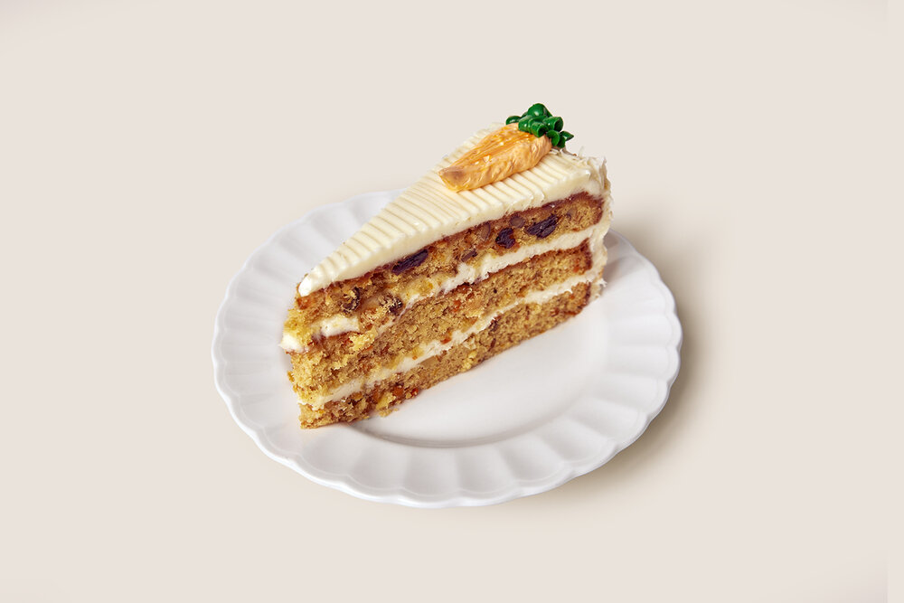 A slice of three-layer carrot cake with cream cheese frosting and a decorative marzipan carrot on top, served on a white scalloped plate.