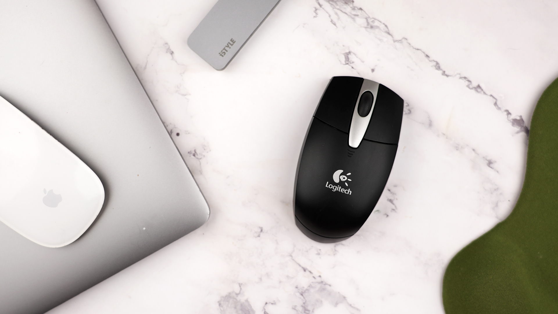 Black Logitech optical mouse beside Apple laptop, white Magic Mouse, and green mousepad on marble desk.