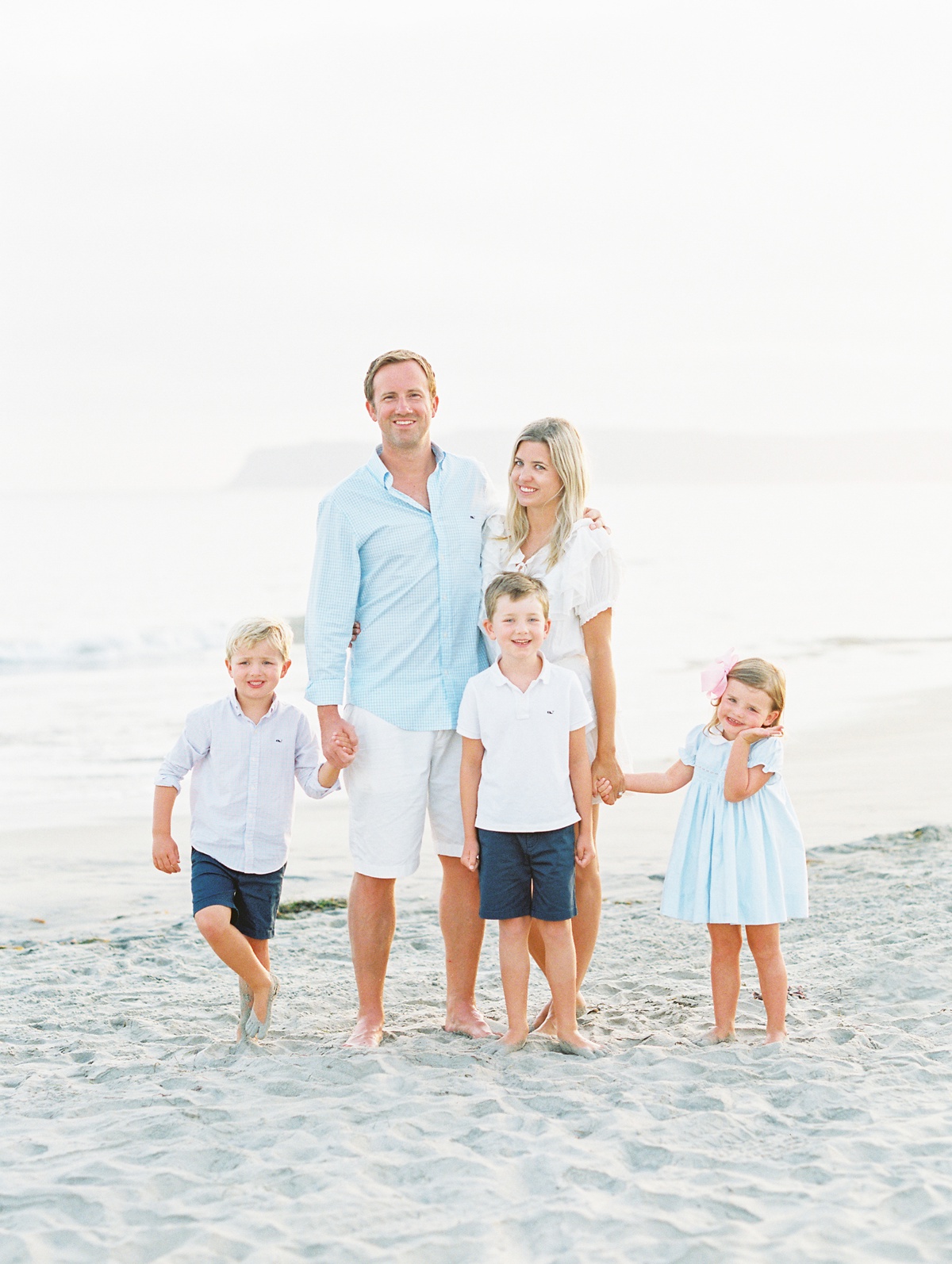 A smiling family of five poses barefoot on a sandy beach at golden hour, dressed in coordinated light blue and white outfits.