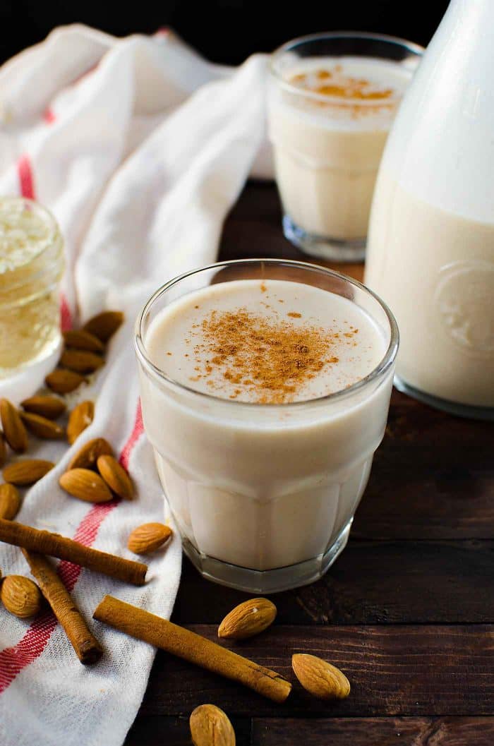 Creamy almond milk drink in a glass, garnished with cinnamon, surrounded by raw almonds and cinnamon sticks on a rustic wooden table.