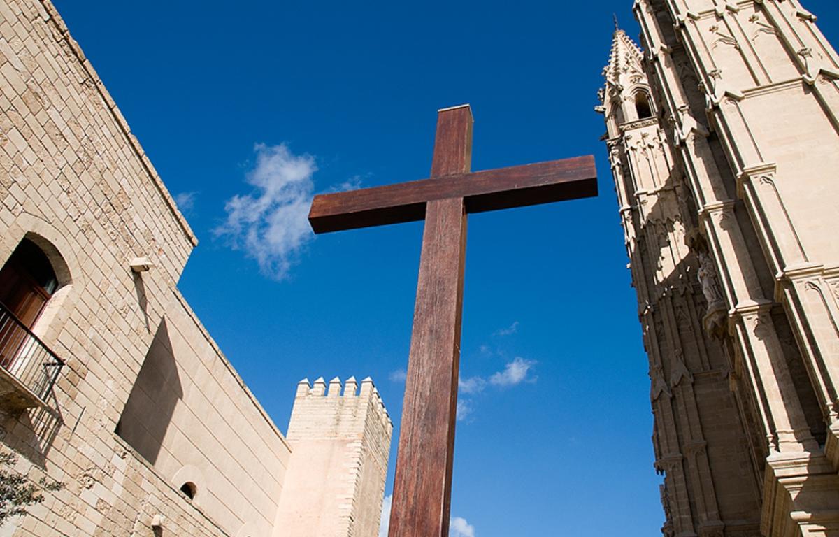 A large wooden cross stands between historic stone buildings under a bright blue sky with scattered clouds.