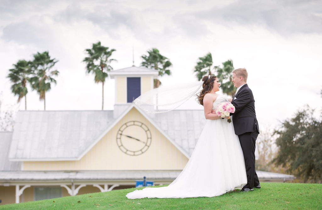 Bride and groom share a joyful moment on a grassy hill with a clock tower and palm trees in the background.