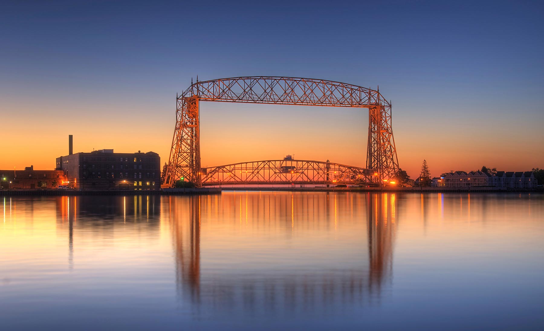 A historic steel lift bridge at sunset, reflected in calm water, with industrial buildings and residential structures on either side.
