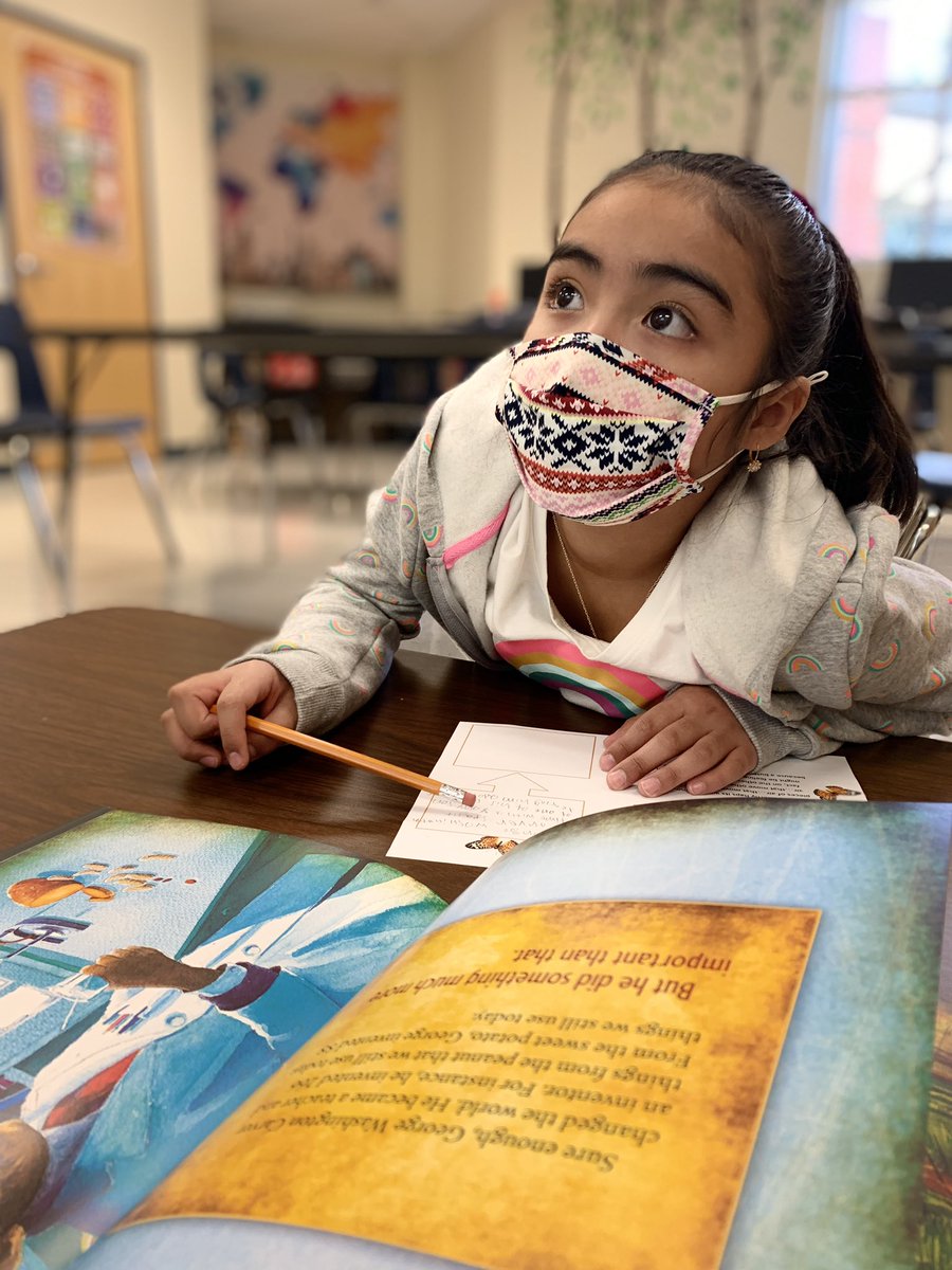 A young girl wearing a patterned face mask reads a colorful book at a classroom desk, holding a pencil and looking upward thoughtfully.