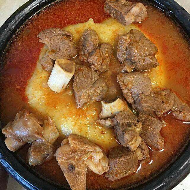 Hearty lamb stew with bone-in chunks and rich red broth served in a black clay pot.