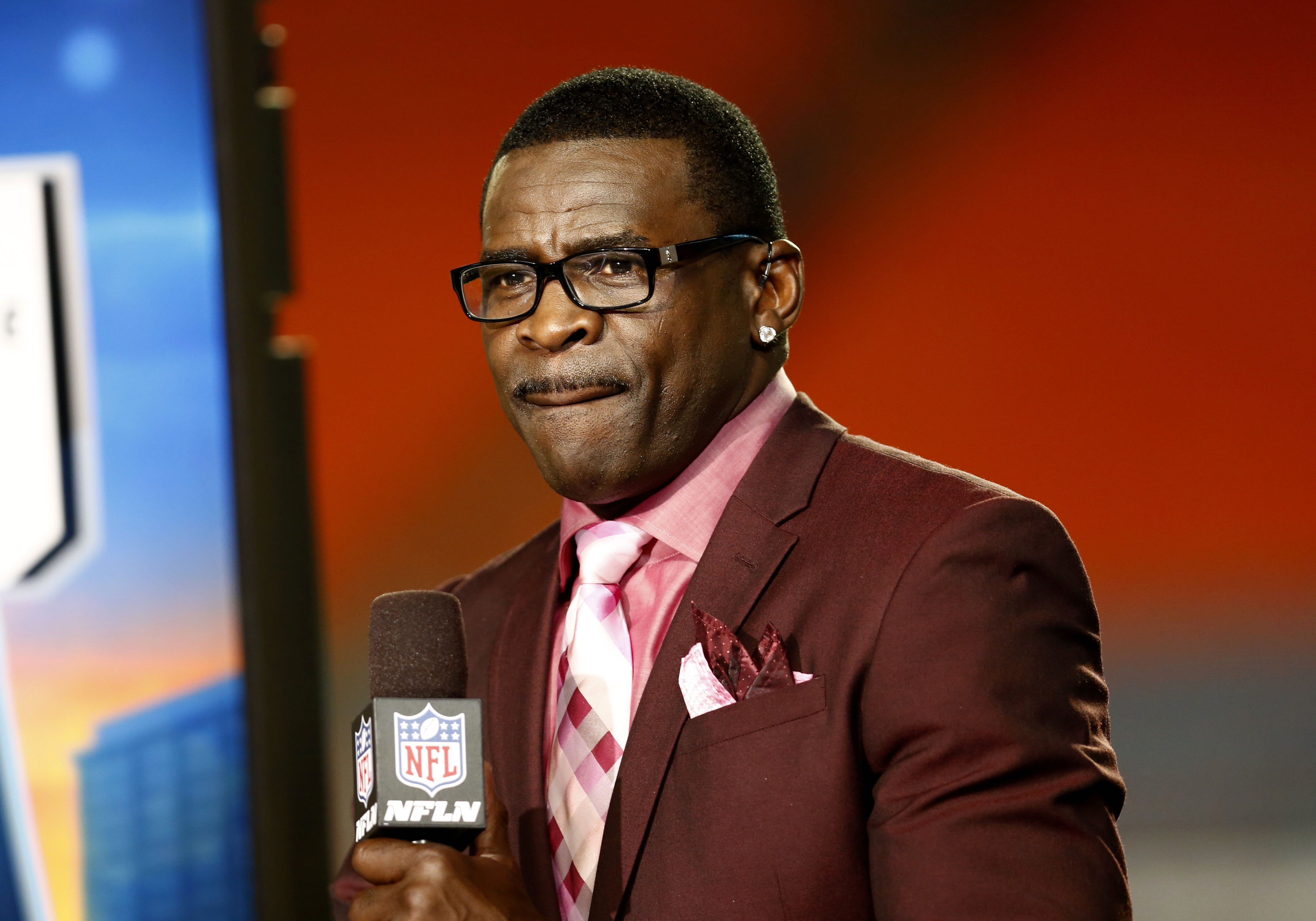 NFL analyst in maroon suit and pink tie holding an NFL Network microphone during a broadcast segment.