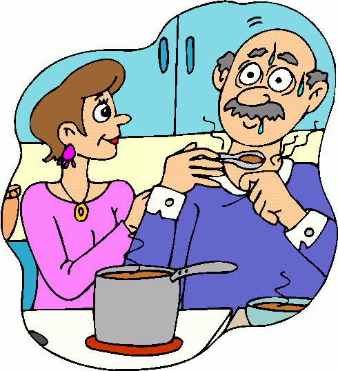 A woman feeds soup to an elderly man with a mustache, who looks surprised and sweaty, in a cozy kitchen setting.