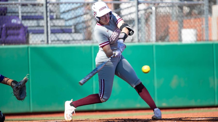 Fordham softball player mid-swing, connecting with a yellow ball during a sunny game.