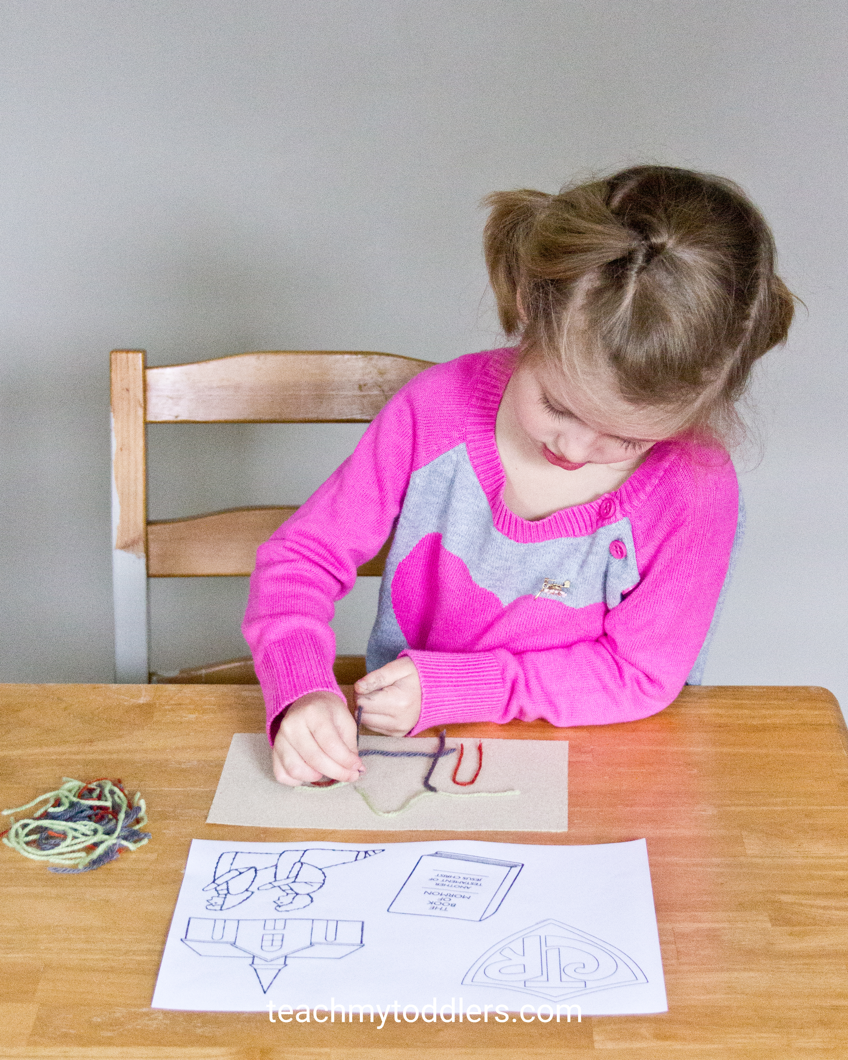 A toddler girl in a pink sweater practices lacing on a felt activity board with yarn, guided by printed templates.