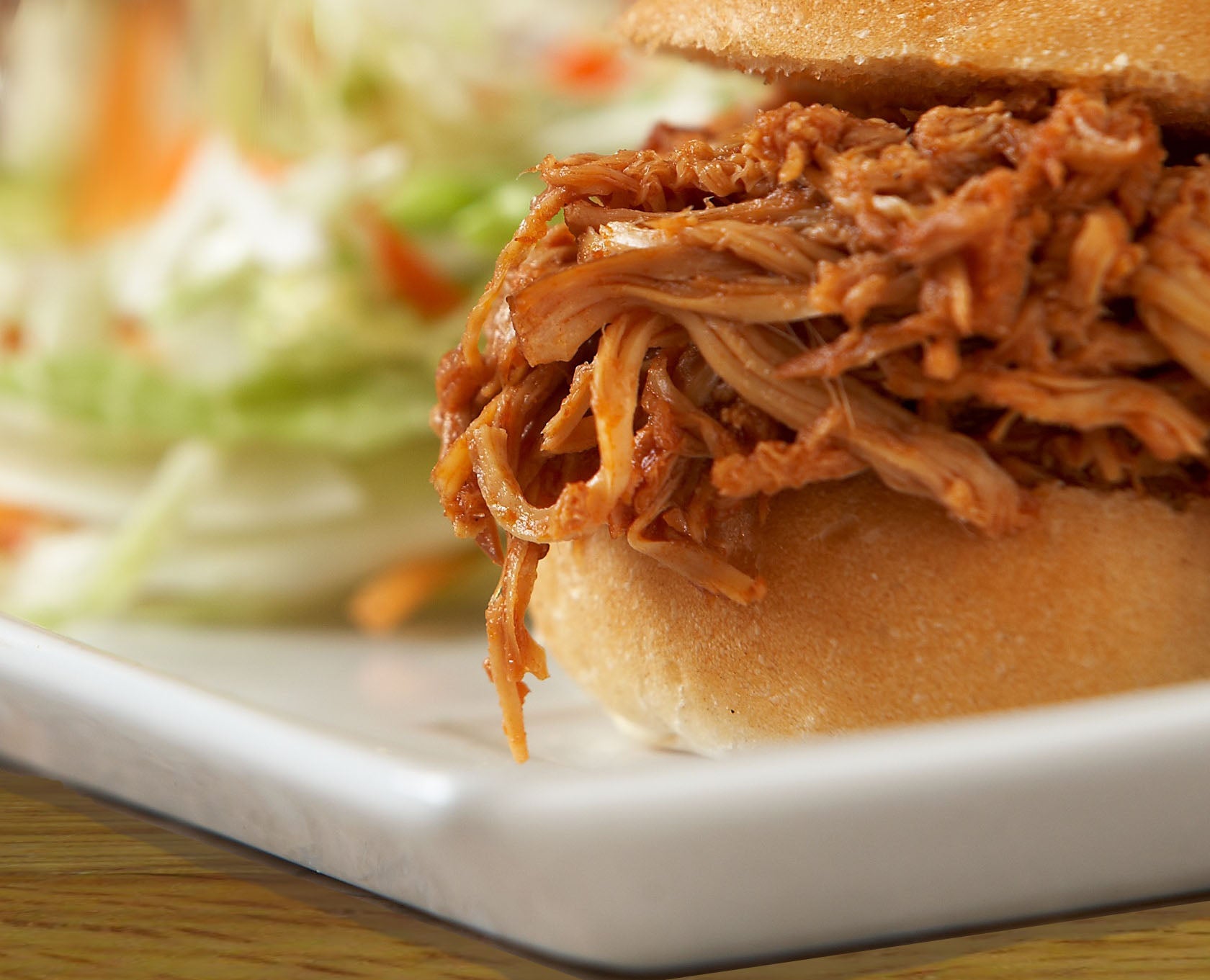 Pulled pork sandwich on a soft bun with coleslaw in background, served on a white plate.