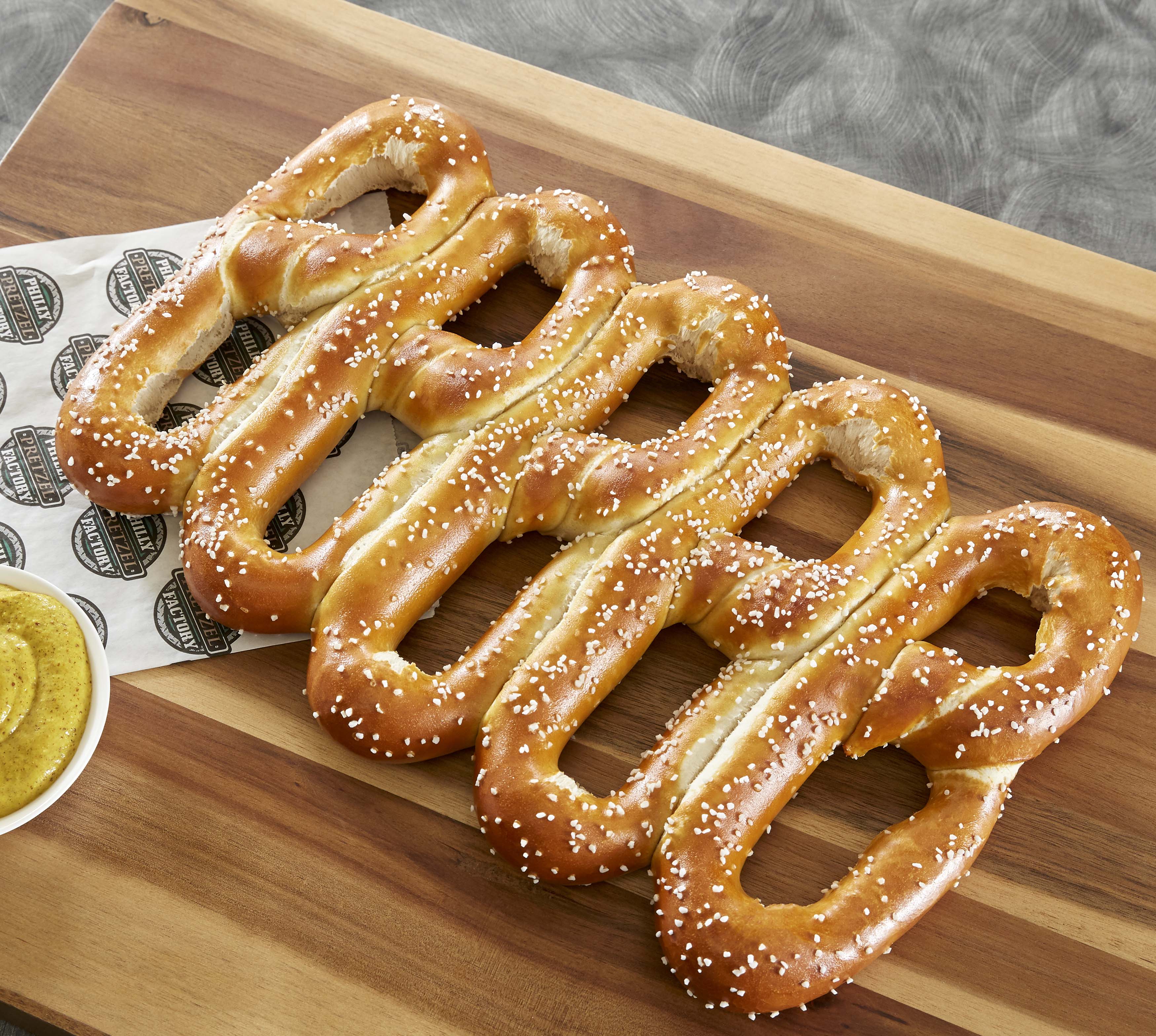 Five golden-brown soft pretzels with coarse salt, served on a wooden board with mustard dipping sauce and branded paper liner.