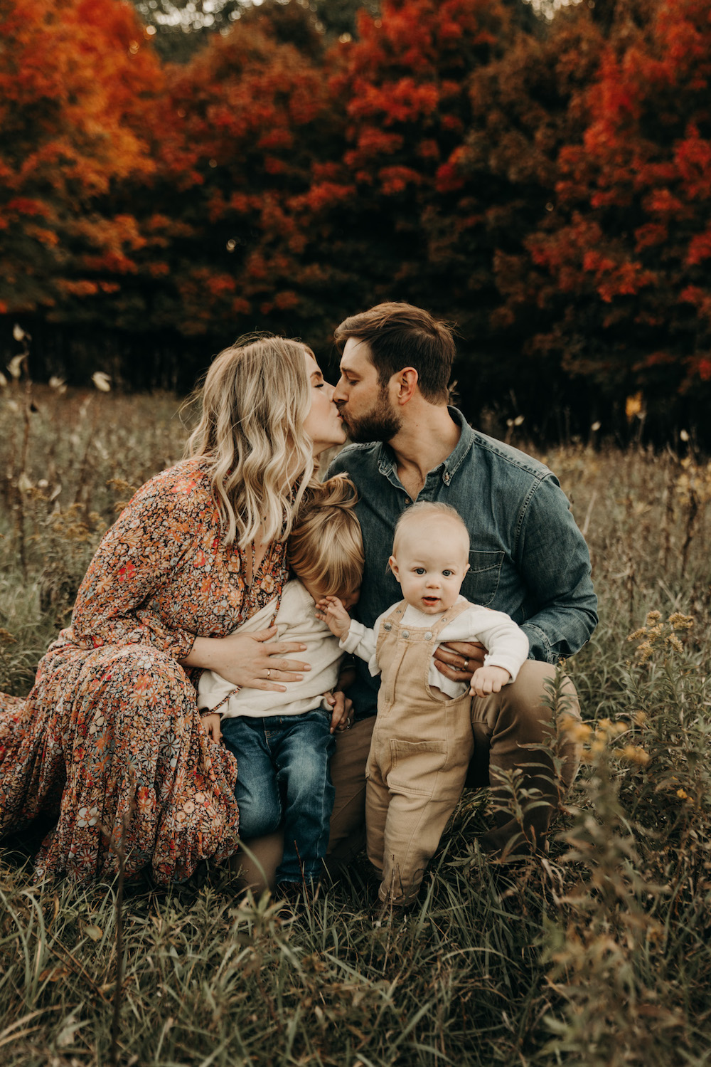 A family of four shares a tender moment in an autumn field, with parents kissing while holding their two young children.