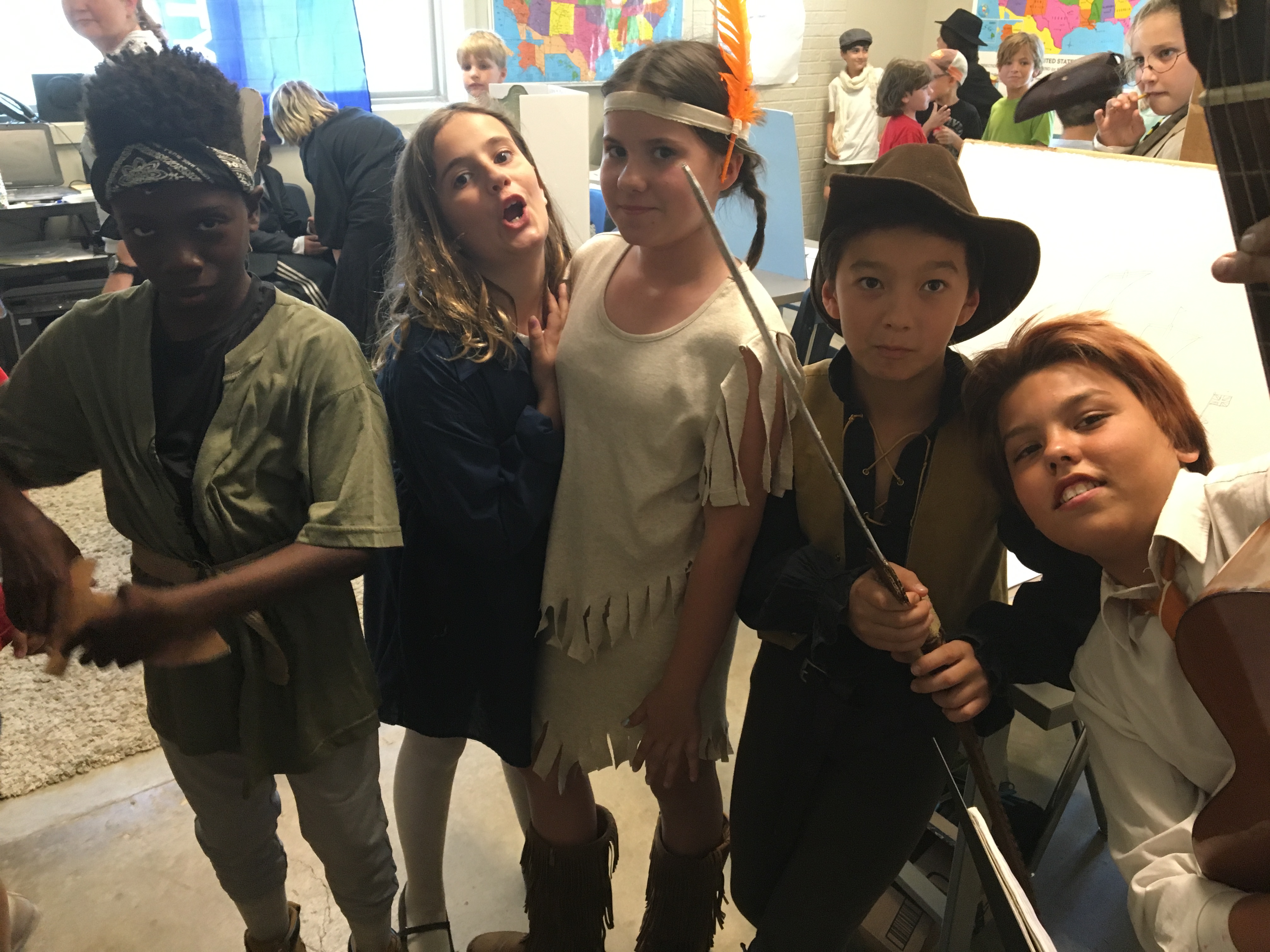 Four children in historical and theatrical costumes posing together in a classroom setting with maps and other students in background.