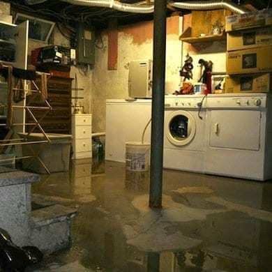 Flooded basement with standing water around washer, dryer, and storage items; signs of water damage on walls and floor.