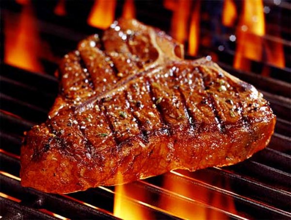 Juicy T-bone steak sizzling on a flaming grill with char marks and herbs.