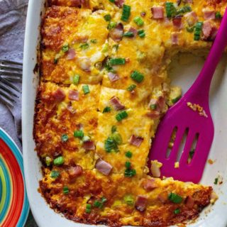Cheesy ham and egg casserole in a white baking dish with a purple spatula, garnished with green onions.