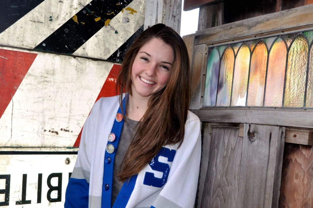 Young woman smiling beside rustic wooden structure with stained glass and warning sign.