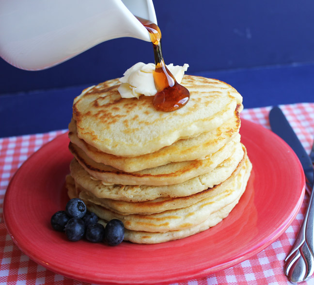 Maple syrup being poured over a stack of golden pancakes with butter and blueberries on a red plate.