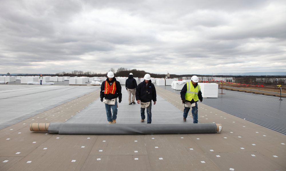 Three construction workers in hard hats and safety vests walk across a flat roof with a large roll of roofing membrane.