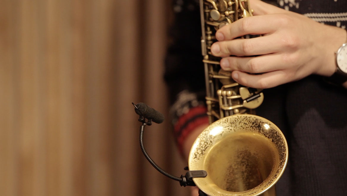 Close-up of hands playing a brass saxophone with a small microphone clipped near the bell, against a warm curtain backdrop.
