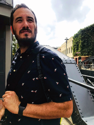 Man with beard and dark patterned shirt standing beside industrial metal structure outdoors.