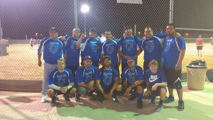 A nighttime group photo of a men's softball team in matching blue "Toros" jerseys posing on a lit field behind a chain-link fence.