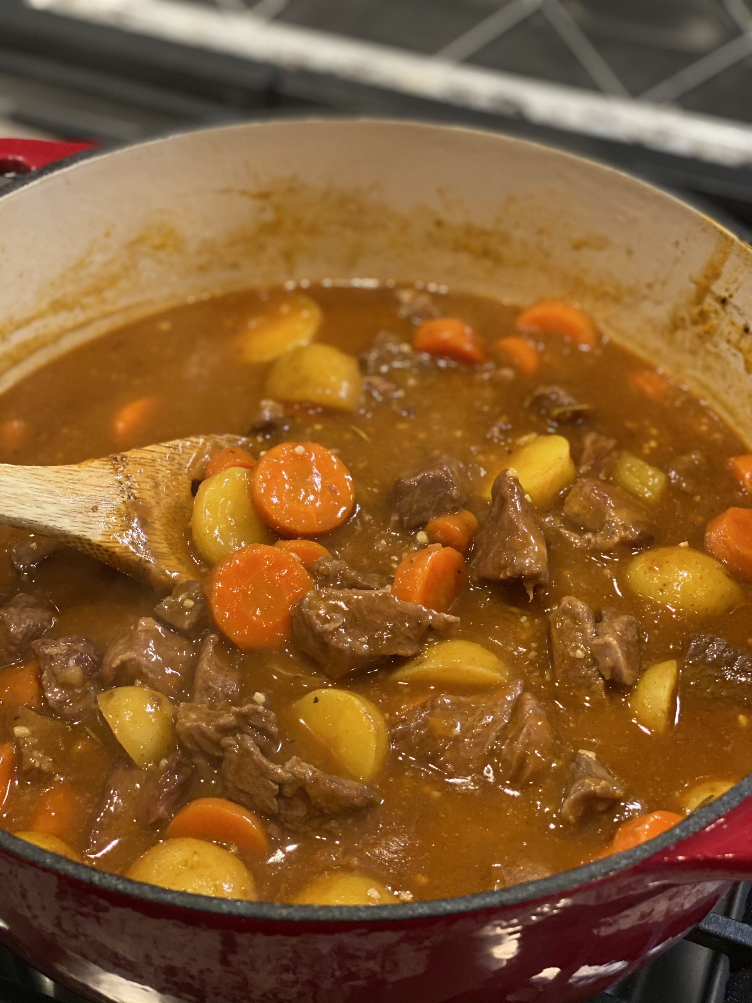 Hearty beef stew with carrots and potatoes simmering in a red Dutch oven on a stovetop.