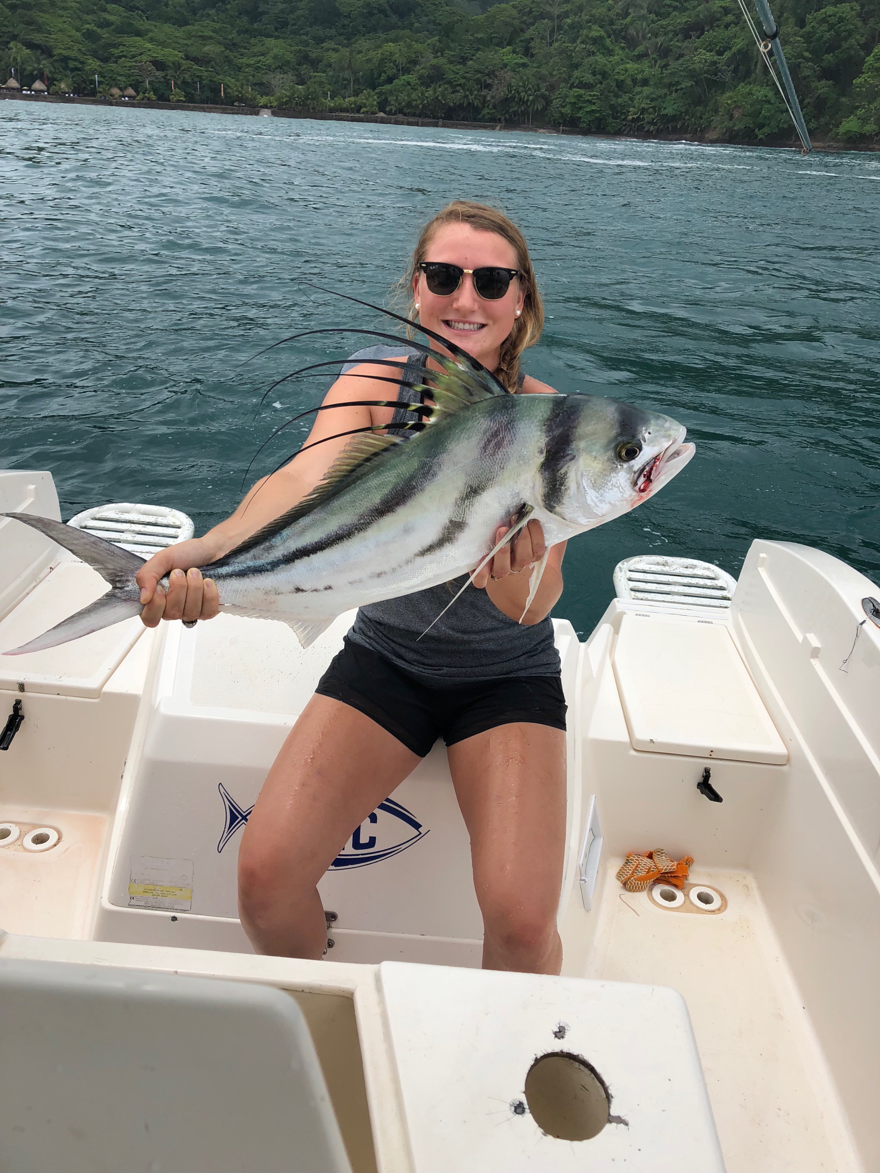 Woman on boat holding a large roosterfish with long dorsal fins, smiling at camera in tropical waters.