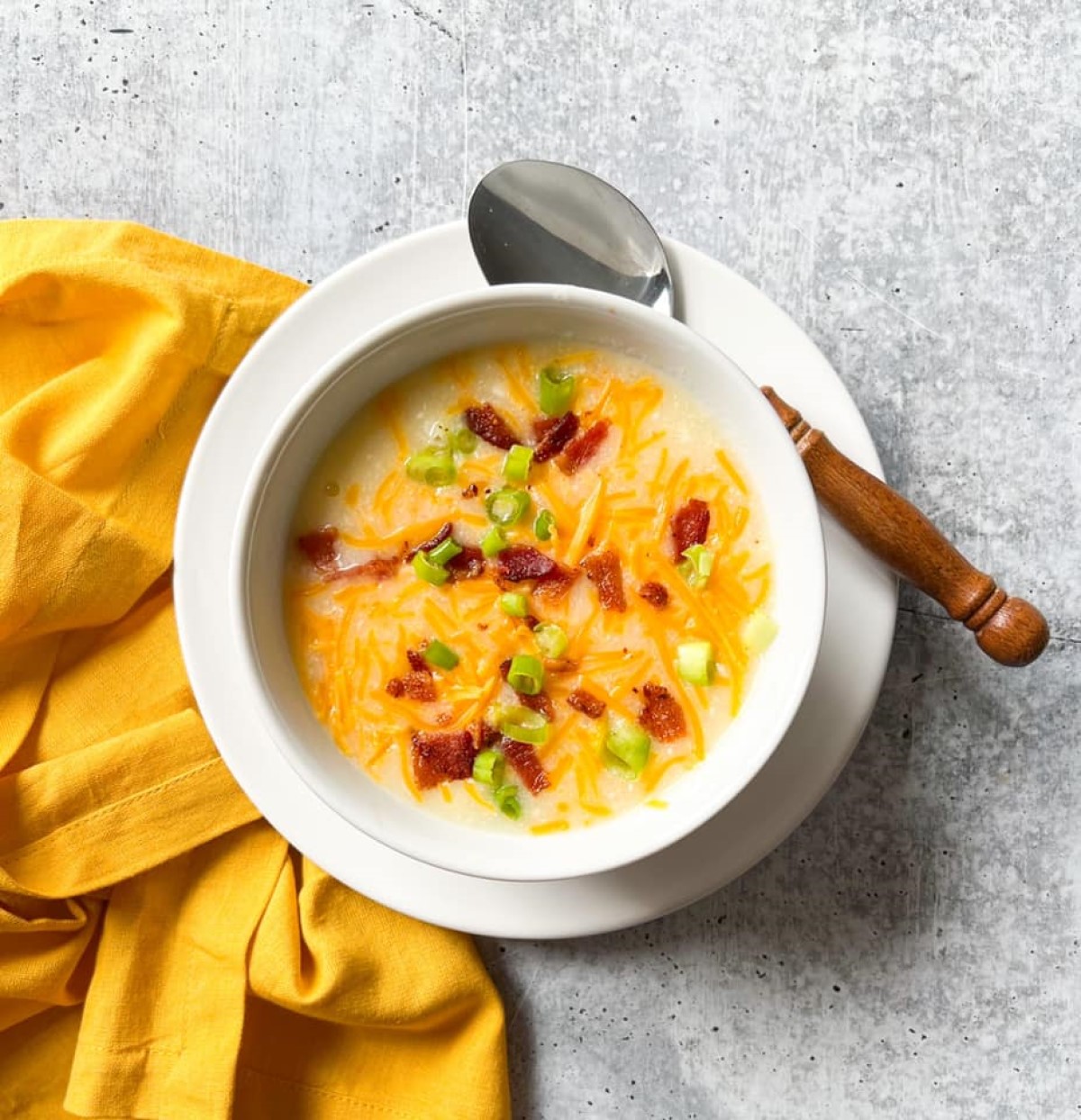 Creamy loaded baked potato soup topped with shredded cheddar, crispy bacon, and green onions in a white bowl with wooden-handled spoon.
