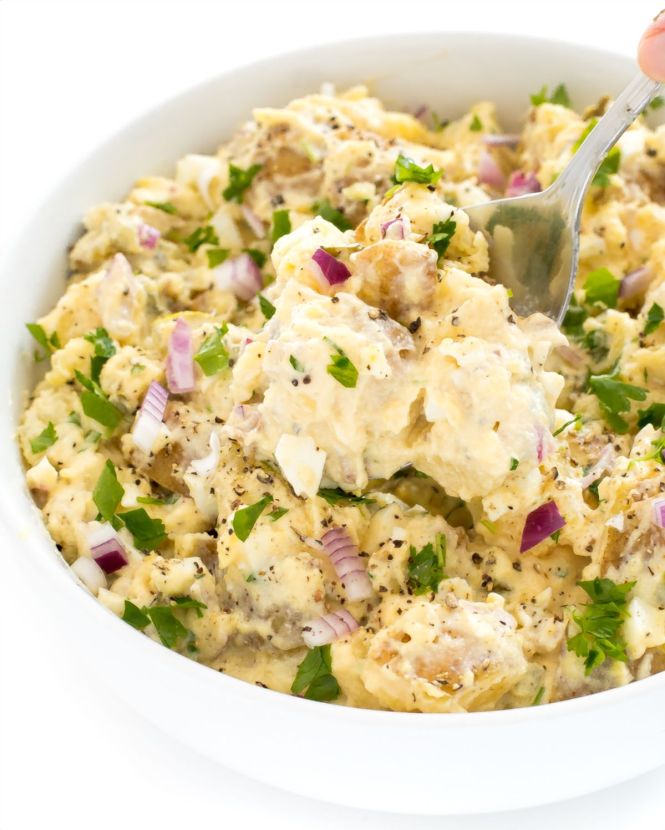 Creamy potato salad with red onion, parsley, and black pepper in a white bowl, spoon lifting a portion.