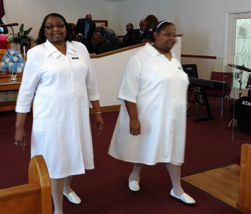 Two women in white choir robes walking down the aisle in a church during a service.