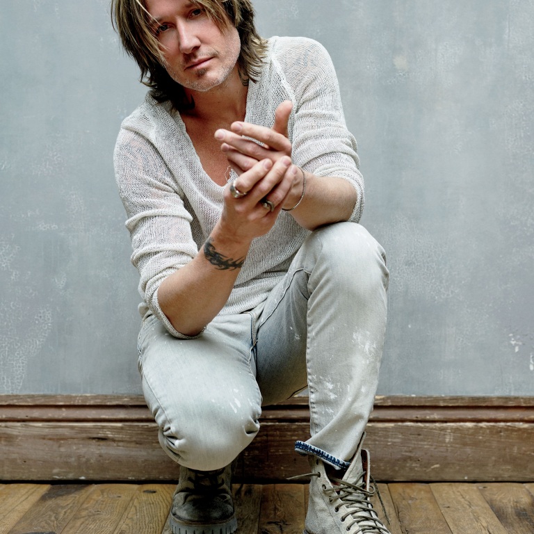 A man with shoulder-length hair crouches on a wooden floor against a gray wall, wearing a light knit sweater and faded jeans.