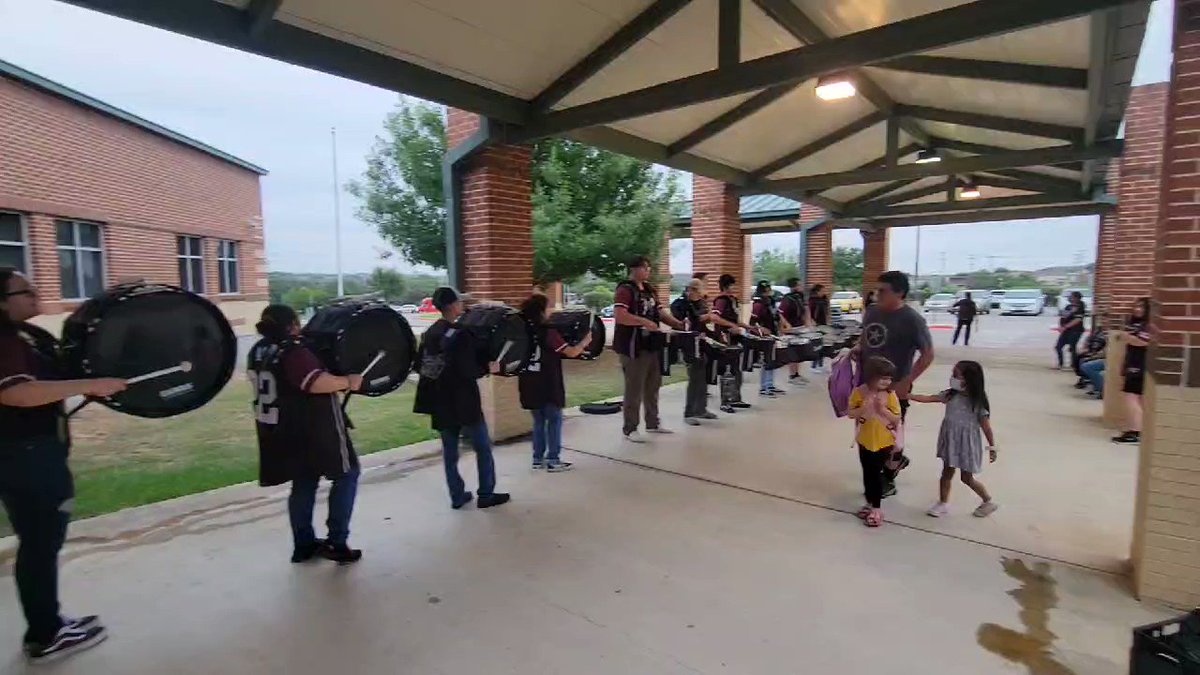School band students practicing under a covered walkway with bass drums and percussion, watched by children and adults.